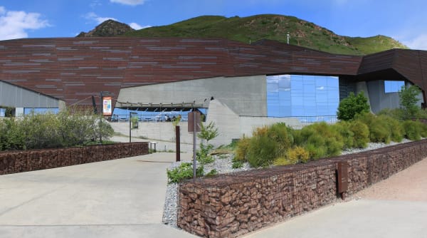 The Natural History Museum of Utah set into the Wasatch foothills — angular copper-clad walls weathered to a dark bronze, a tall glass curtain wall reflecting cloud, and board-formed concrete beneath. Gabion retaining walls of stacked rock and beds of native grasses line the approach path, and the green hillside rises directly behind the roofline.