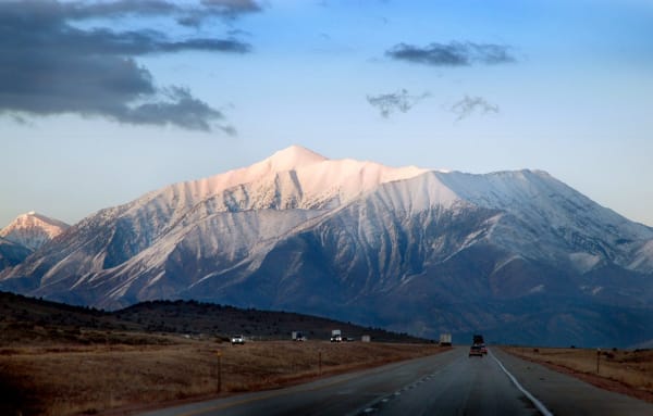Mount Nebo under fresh snow seen head-on from the highway at valley level — the peak's long snow-covered ridge catching low pink light above dark timbered foothills, with the road running straight toward it in the foreground and a car and several semi-trailers strung out along the lanes.