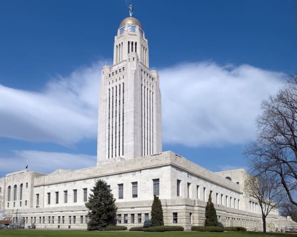 The white limestone tower of the Nebraska State Capitol, topped by a gold dome, rises above its square base under a blue sky in Lincoln.