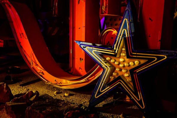 Salvaged signs in the Neon Museum's boneyard — a large steel star faced with round gold bulbs and outlined in blue, standing against the curved orange sweep of another sign, on a bed of gravel and broken rock.