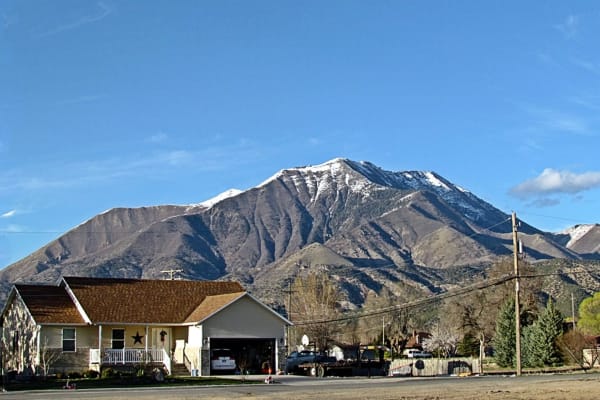 A cream colored house with a brown shingled roof and a white porch rail on a quiet residential street, an open garage beside it and a flatbed truck at the curb. Behind the rooftops a long ridged mountain rises, its summit and upper slopes streaked with snow.