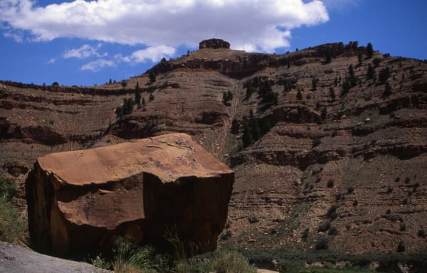 A large fallen block of red sandstone lying beside the gravel road on the floor of Nine Mile Canyon, its top face weathered smooth and its broken sides in deep shadow. The canyon wall rises behind in stepped ledges of red-brown rock with scattered junipers and a capped butte on the skyline, under a blue sky with white cloud. No rock art is visible in the frame.