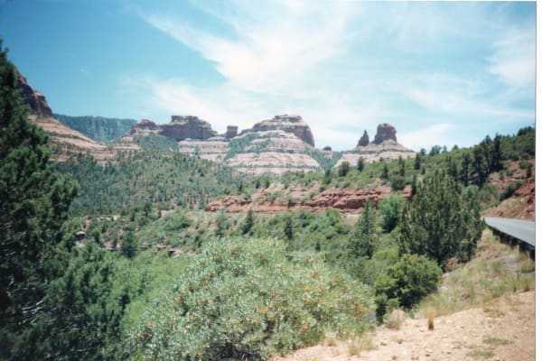 Oak Creek Canyon looking up-canyon on faded colour film — banded buttes of red rock over pale cream closing the head of the canyon, dense ponderosa and juniper covering both walls, and a red ledge running across the middle distance. State Route 89A shows only as a short length of steel guardrail at the right edge, and none of the switchbacks the drive is known for is in the frame. The near foreground is swept into horizontal blur by camera motion while the far canyon stays sharp.