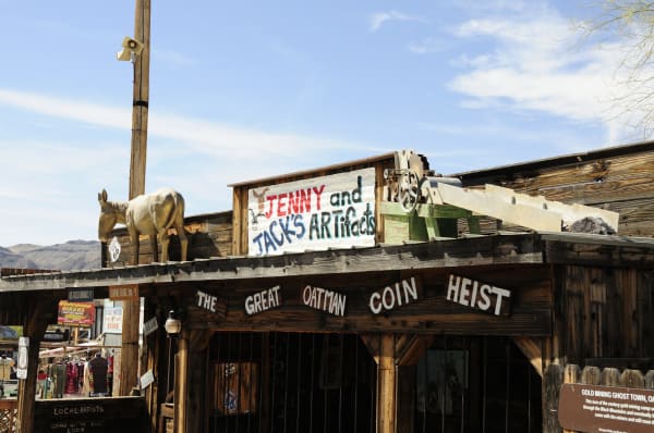 Storefronts on the main street of Oatman shot from below the eaves — a model burro standing on the porch roof at the left beside a Route 66 shield, a hand-lettered board reading JENNY and JACK'S ARTifacts behind it, and a piece of old ore-processing machinery with a green frame and a chute set along the roofline. A heavy timber beam across the front is painted THE GREAT OATMAN COIN HEIST, and the shopfronts beneath carry hand-painted signs for ice-cold drinks, burro food, leather goods and local artists. An interpretive panel headed GOLD MINING GHOST TOWN stands at the right. The live burros that roam the street, the Black Mountains and the Sitgreaves Pass switchbacks are all out of frame.