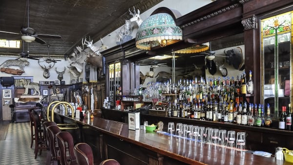 The long wooden bar of the Occidental Hotel saloon in Buffalo, its back bar lined with bottles under a stained-glass lamp and mounted heads on the walls