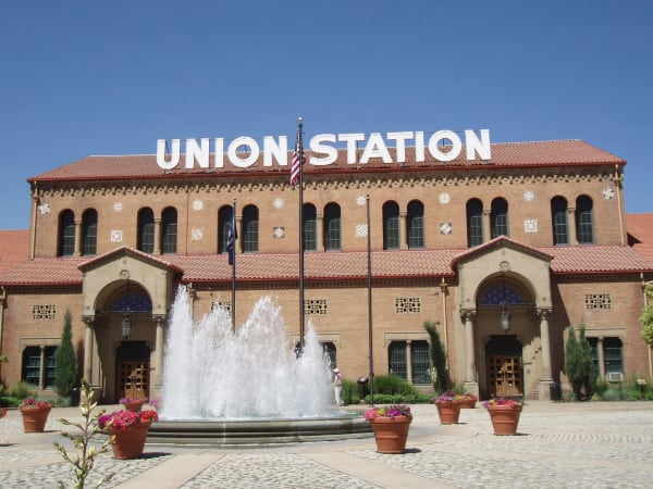 Ogden Union Station's long brick facade beneath a red tile roof, with UNION STATION spelled out in large white letters along the roofline, rows of tall arched windows set between coloured tile medallions, and twin arched entrance porches carried on stone columns. A fountain plays in the paved plaza in front, ringed by terracotta pots of flowers, with the US and Utah flags on poles alongside.