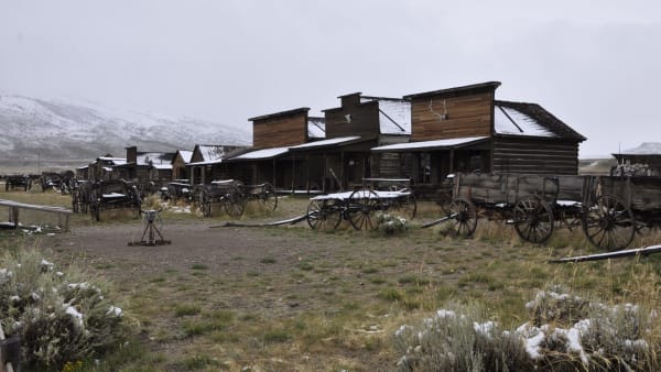 A row of old false-fronted log buildings at Old Trail Town in Cody, wagons parked in front and a dusting of snow on the hills