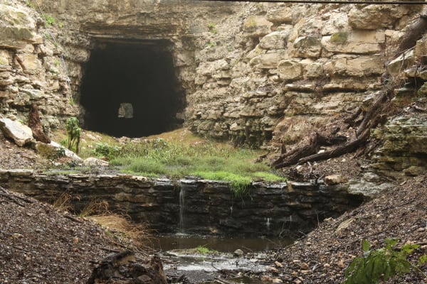 The dark arched mouth of the old railroad tunnel in a cut of layered limestone at Old Tunnel State Park, with a small pool below.
