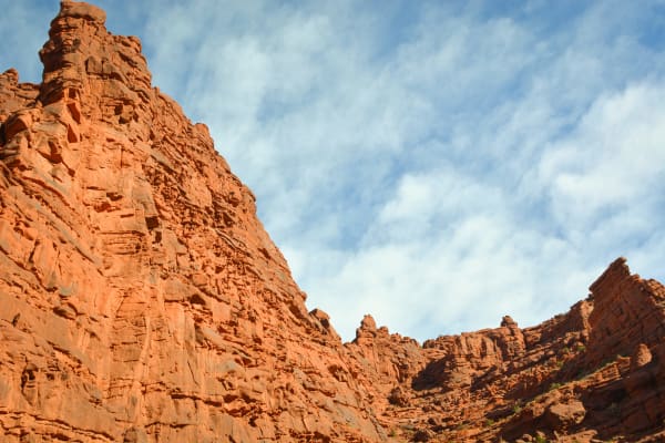 Steep orange-red canyon walls seen from below against a blue sky streaked with high cloud, a buttressed cliff filling the left of the frame and lower broken ledges stepping away to the right. Sparse green brush grows along the rims.