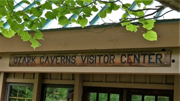 A carved wooden sign reading Ozark Caverns Visitor Center over the entrance, framed by summer leaves.