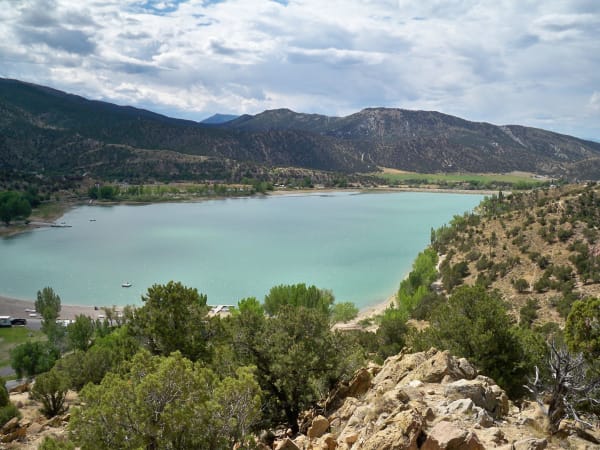 A turquoise reservoir seen from a juniper covered bluff above it, a pale beach curving along the near shore, a few small boats out on the water and vehicles parked at a day use area. Wooded hills ring the far side and broken cloud fills the sky.
