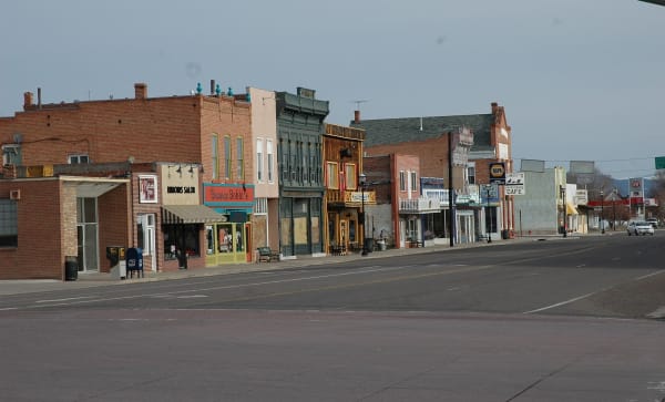 A row of Panguitch's brick Main Street storefronts along an empty, unusually wide street — red and painted brick fronts with parapets and false fronts, a green-painted cast-iron shopfront at centre, a striped awning over a salon, a red sign reading Bronco Bobbi's beside it, and further signs for a cafe and an auto parts store down the block. A mail box, a bench and black lamp standards stand on the sidewalk, bare trees line the far end, and a low range shows in the distance under flat grey sky.
