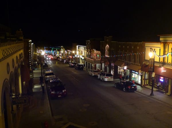 A historic main street at night seen from above, strings of white and colored lights along the storefronts, lit shop windows and neon signs, cars parked at both curbs and a line of red taillights receding uphill into the dark.