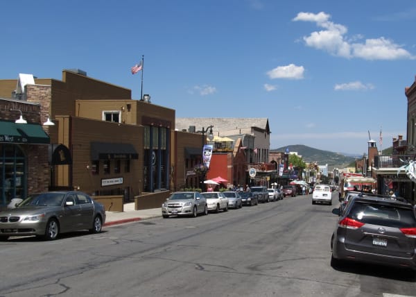 A historic mining-town main street in summer, looking down the roadway between rows of parked cars, low nineteenth-century storefronts with awnings and patio umbrellas on both sides, a US flag on a pole, and a green hillside closing the view at the end of the street.