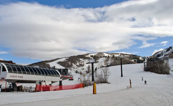 The base of a beginner ski run in winter, a covered chairlift with a glazed hood at the left signed FIRST TIME, orange safety netting across the snow, lift towers marching up the slope behind and a few skiers on the groomed run. Forested ridges rise beyond under heavy cumulus.