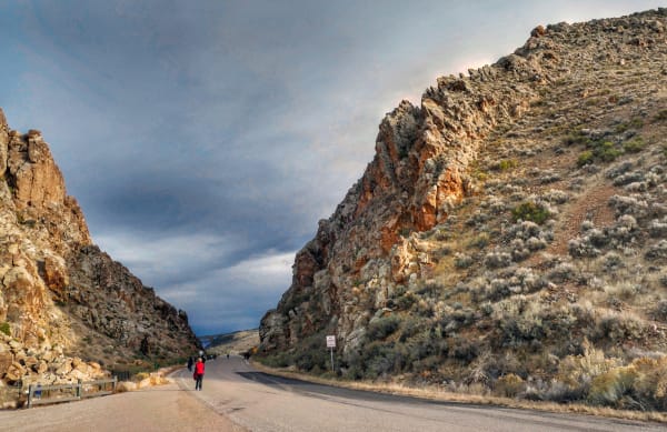 Parowan Gap — the notch an old river cut through the Red Hills — seen from the pavement that runs through it, rough tan and grey rock rising steeply on both sides above sagebrush slopes with a bank of grey cloud filling the sky. A group of people walks up the road through the gap, the nearest in a red vest with her back to the camera, and a yellow pedestrian crossing sign stands at the right. None of the petroglyph panels are legible in the frame.