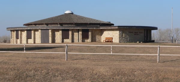 The round limestone Pawnee Indian Museum with its low shingled roof, seen across a winter pasture behind a white rail fence.