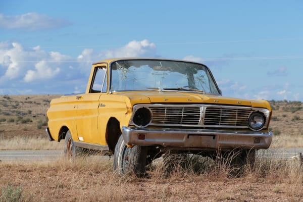 A derelict yellow Ford with a low open cargo bed standing in dry roadside grass near Peach Springs, filling the frame in three-quarter view. Its windscreen is crazed from edge to edge, one headlight is missing from its bezel, the panels and wheel arches are rusting through, and the near wheel is a bare rim with a rotted tyre still on it; FORD is lettered in chrome across the front edge above the grille. A strip of pavement runs behind it and open scrub reaches away to low ground under a blue sky with cumulus. Peach Springs itself is not in the frame: no part of the town, none of the Hualapai tribal government buildings, and no view to the canyon rim.