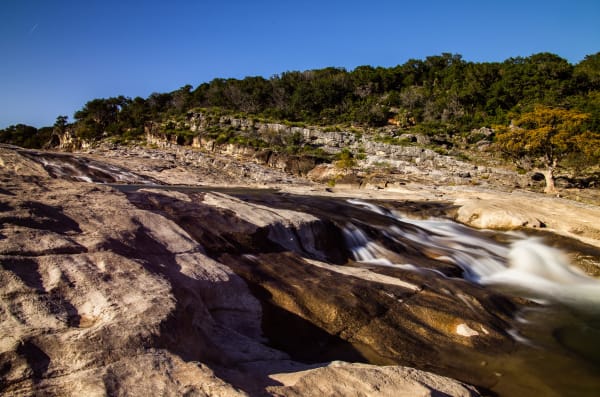 The Pedernales River spilling over smooth layered limestone at Pedernales Falls, with a wooded bluff behind.