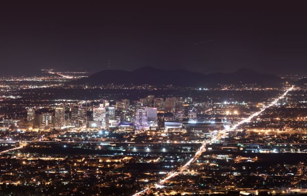 Phoenix at night from high ground to the south. The downtown towers stand lit in a compact cluster left of centre with an arena beside them, and the lit street grid runs out flat to the horizon in every direction. A major arterial cuts diagonally across the right of the frame as an unbroken line of white and amber, low ridges show dark against the glow along the skyline, and the sky above is empty and starless.