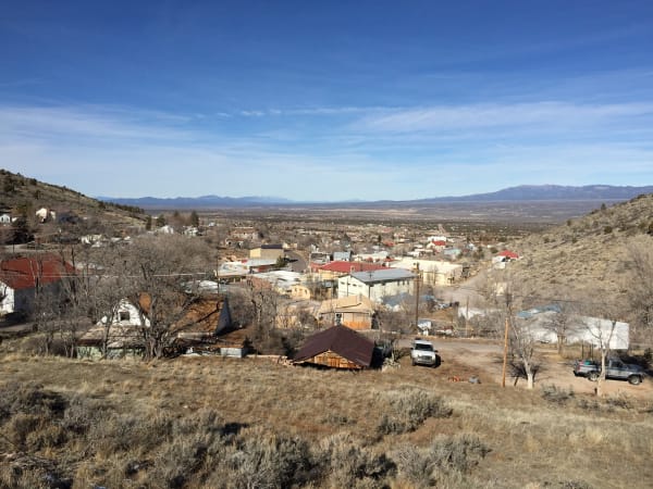 Pioche seen from the hillside above, the town running down the slope and out onto the valley floor — tin and shingle roofs, a red-roofed commercial block and a large pale two-storey building along the curve of the main street, houses and sheds among bare winter trees, and pickups parked on dirt turnouts in the near ground. Dry grass and sagebrush cover the foreground slope, and the valley runs flat and pale to a long snow-touched range on the horizon. Boot Hill, the courthouse and the aerial tramway are not identifiable in the frame.