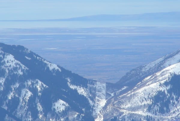 A hazy long-lens view from high on Powder Mountain, looking down a snow-covered canyon between conifer-studded slopes to the valley floor opening out below, with a road threading the canyon bottom at right and pale flats fading into haze beneath a long low range on the horizon.
