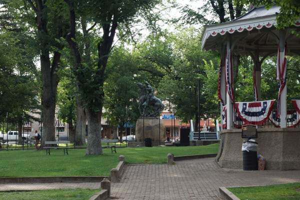 The Yavapai County Courthouse Plaza at Prescott under big shade elms — the Rough Rider Monument at the centre, Solon Borglum's 1907 bronze of a mounted man on a plunging horse set on a rough granite boulder, and the plaza bandstand at the right hung with red, white and blue bunting. Picnic tables and a brick path cross the grass, one walker passes at the left, and storefronts stand across the road behind under striped awnings. The courthouse building itself is out of frame.