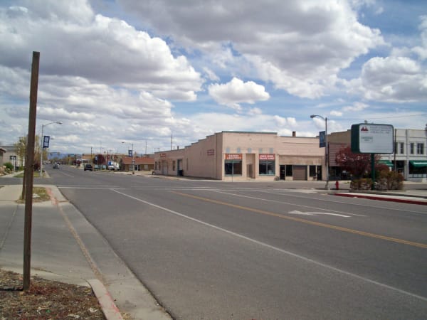 Price's main street on a bright day with cloud filling most of the frame, looking down a wide empty four-lane road past a bare timber utility pole in the near foreground. A low stuccoed commercial block stands along the right side, orange signs above two of the shopfronts reading THINGS AND STUFF and PRICE RIVER VALLEY LOAN, and a green board for a community health office on a post beyond, with bare trees and further storefronts running away down the block.