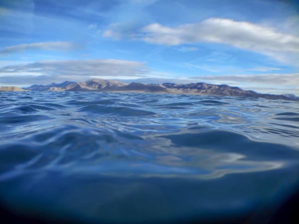 A view from just above the water surface at Pyramid Lake, low waves running toward the camera and a long bare desert range rising along the far shore under a blue sky with high cloud.