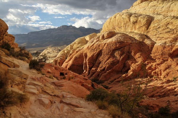 Looking up a wash in Red Rock Canyon from a sandy trail in the foreground, humped red sandstone filling the middle of the frame with pale cream rock capping it at the upper right, a dark mountain range across the valley behind and storm cloud overhead.