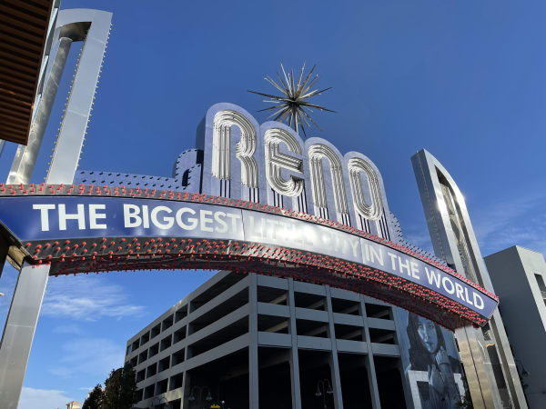 The Reno arch spanning Virginia Street by day, its lettering reading "RENO" and "THE BIGGEST LITTLE CITY IN THE WORLD" beneath a starburst, with a concrete parking structure behind it under a clear blue sky.