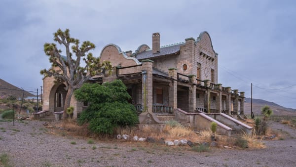 The 1908 Las Vegas and Tonopah Railroad depot at Rhyolite, a Mission Revival building of pale rock-faced concrete block with shaped curvilinear gables, green-trimmed parapets, a brick chimney and a deep arcaded veranda along its front, its window openings boarded or empty. A Joshua tree stands against the left gable and a large green shrub has grown across the porch steps; gravel, dry grass and scattered stone fill the foreground, and bare hills close the view under heavy overcast. The Cook Bank ruin and the opera house are not in the frame.