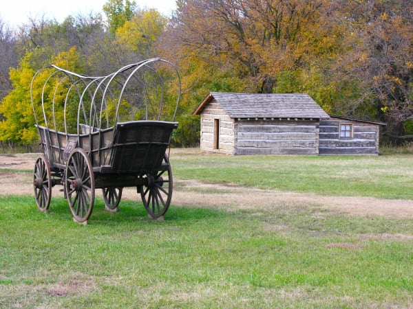 A wooden wagon with bare bows stands on the grass in front of a log building at Rock Creek Station, with autumn trees behind.