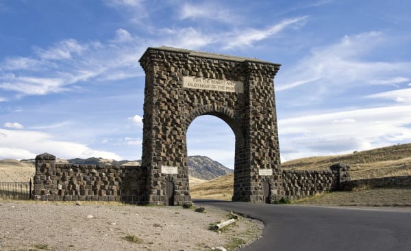 The Roosevelt Arch at Gardiner, a tall arch of dark basalt blocks with low wing walls, the road passing through under a blue sky