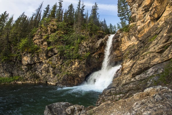 Running Eagle Falls pouring through a notch in a tan rock wall into a dark pool, firs along the cliff top