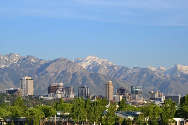 Downtown Salt Lake City's towers seen across a belt of green treetops, with the Wasatch Range rising directly behind the city and snow still lying on the high peaks and ridgelines under a clear blue sky. The white spires of the Salt Lake Temple stand among the office blocks left of centre.