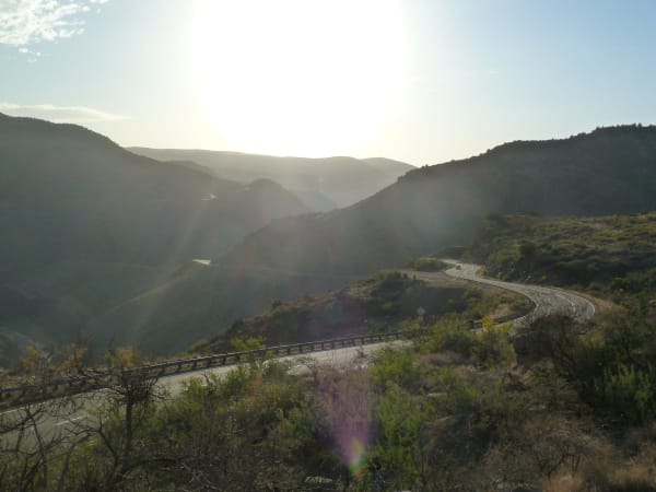 US-60 descending into Salt River Canyon, shot into a low sun with heavy lens flare across the frame — the highway curving through a hairpin with guardrail along its outer edge and a single pale pickup on the bend, another length of the road visible against the far wall. Brushy slopes fall away on both sides and hazy ridges close the canyon behind. The Salt River itself, and the boundary it draws between the White Mountain and San Carlos Apache nations, are not visible in the frame.