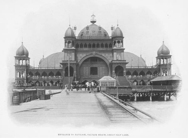 An archival photograph captioned "Entrance to Pavilion, Saltair Beach, Great Salt Lake" — the Moorish-domed resort pavilion raised on timber pilings above the water, with onion-domed corner towers, tiers of fretwork verandas, and a railway track running out along the plank pier toward its arched entrance, where a scattering of people in period dress stand.