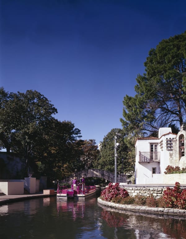 A bend of the San Antonio River Walk with flower beds, stone walls and a white Spanish-style building above the water.