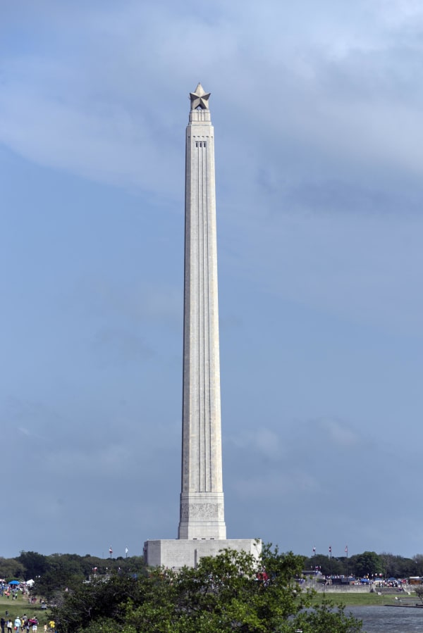 The San Jacinto Monument, a tapering stone shaft topped by a star, rising above trees against a blue sky.