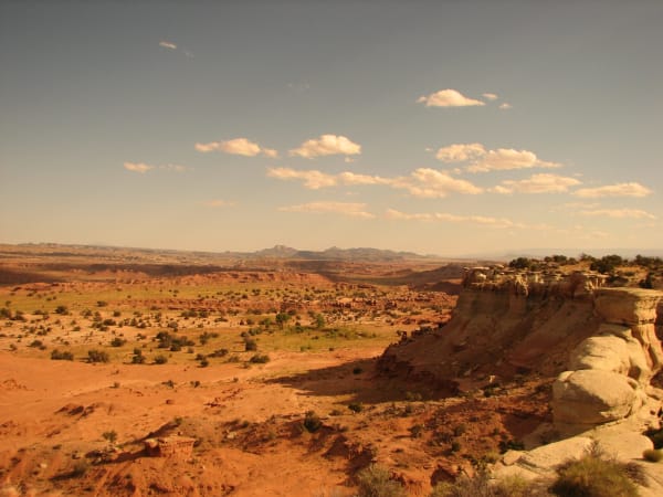 A broad red desert basin in the San Rafael Swell under warm low light, sagebrush scattered across bare red sand and slickrock in the foreground and a low banded escarpment cutting across the middle distance. A white-capped sandstone bluff stands at the right edge and pale reefs run back to a hazy horizon.