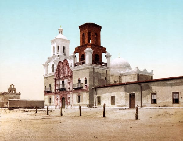 A colour print of Mission San Xavier del Bac issued by the Detroit Photographic Company and copyrighted 1902, the church seen obliquely across a bare sand plaza with wooden posts standing in it. The carved Baroque portal rises in red-brown between the towers, statues set in its niches and turned-wood balconies above the door; the west tower is whitewashed under a domed lantern, and the east tower beside it stands in bare unplastered brick, never finished. A ruined bell gable shows behind a wall at the left. The lantern in this frame is the eighteenth-century original, destroyed by lightning in 1939.