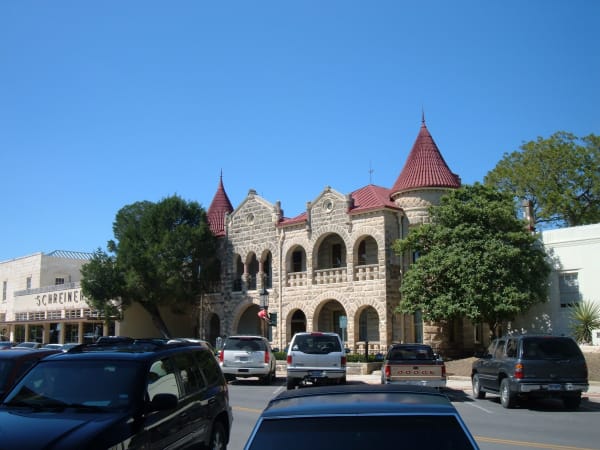 The stone Schreiner Mansion in downtown Kerrville, with arched galleries and red conical turret roofs, behind parked cars.