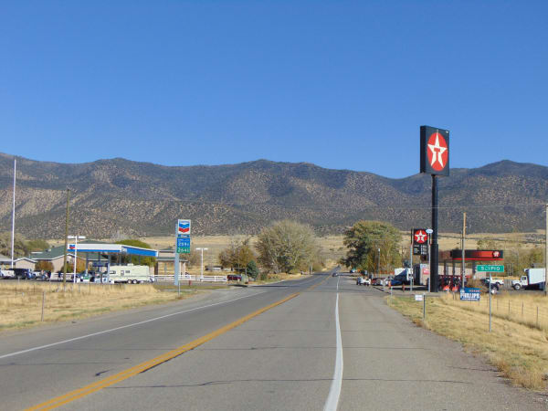 A two-lane highway running straight toward low brown foothills, photographed from the empty roadway. A Chevron station with a blue and white canopy stands on the left behind a white rail fence, and a tall Texaco sign on a black pole marks a second station and store on the right.