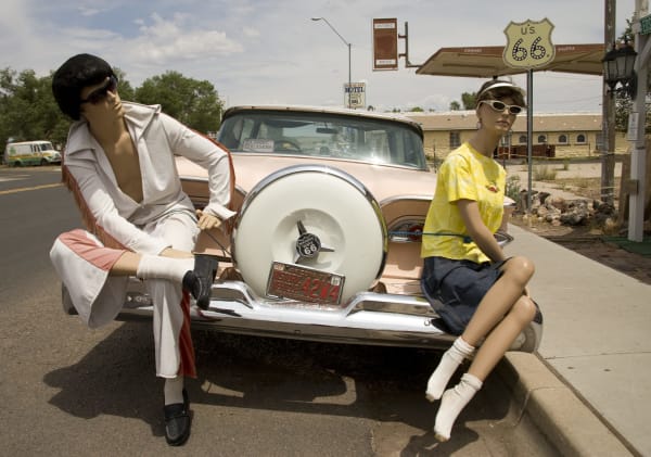 Two shop mannequins posed on the rear bumper of a pink-and-cream 1950s Ford parked at the kerb in Seligman — one dressed as Elvis in a white fringed jumpsuit, dark wig and sunglasses, leaning back against the continental spare-wheel cover, the other in a yellow t-shirt, denim skirt and white visor sitting on the fender with her legs crossed. A CLASSIC CRUISERS decal is on the rear window and an Arizona historic-vehicle plate on the spare cover. Behind them a tall U.S. 66 shield stands on a post beside an awning lettered CHICAGO and SANTA MONICA, with a ROUTE 66 MOTEL sign and a low stone building with arched windows beyond. Angel Delgadillo's barbershop is not in the frame.
