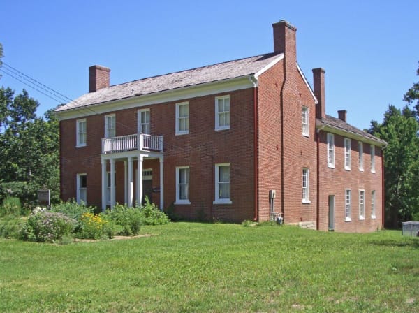 The two-story red brick West Building of the Shawnee Indian Mission in Fairway, with a small columned porch, on a summer lawn.