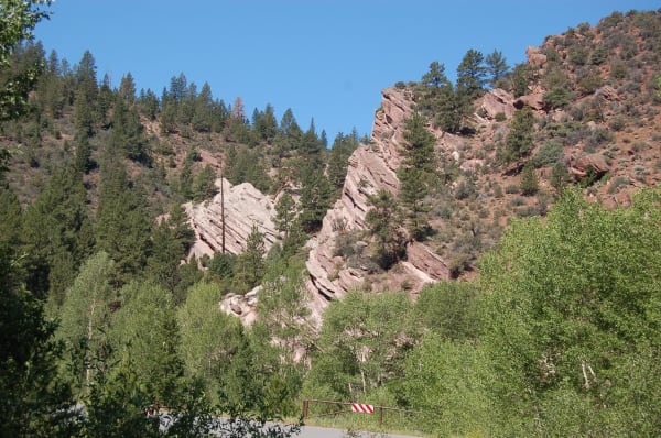 Steeply tilted beds of red sandstone in Sheep Creek Canyon, two fins of rock leaning at a sharp angle out of a conifer-covered slope with their layers stacked visibly along the tilt. Aspens and cottonwoods crowd the foreground, and a brown steel gate carrying a red and white chevron panel stands across the paved road at the bottom of the frame.