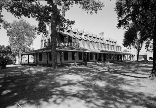 A black-and-white survey photograph of the Sheridan Inn, a long porch-wrapped building with a row of gabled dormers, shaded by big cottonwoods