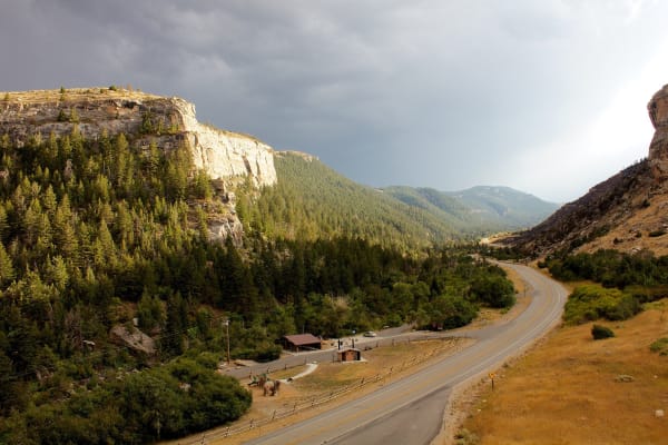 Sinks Canyon seen from its mouth, a road winding up between pine slopes beneath pale limestone cliffs under a stormy sky