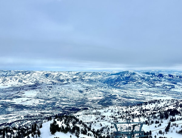 A wide view from high on Snowbasin looking out over range after range of snow-covered ridges and valleys running back to a hazy horizon, with groomed runs and scattered conifers dropping away below and the steel head of a lift terminal standing in the near corner, under flat grey winter cloud.